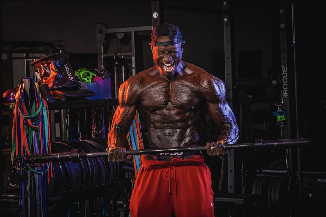 Coach JR curling a barbell in a dark gym lined with resistance bands