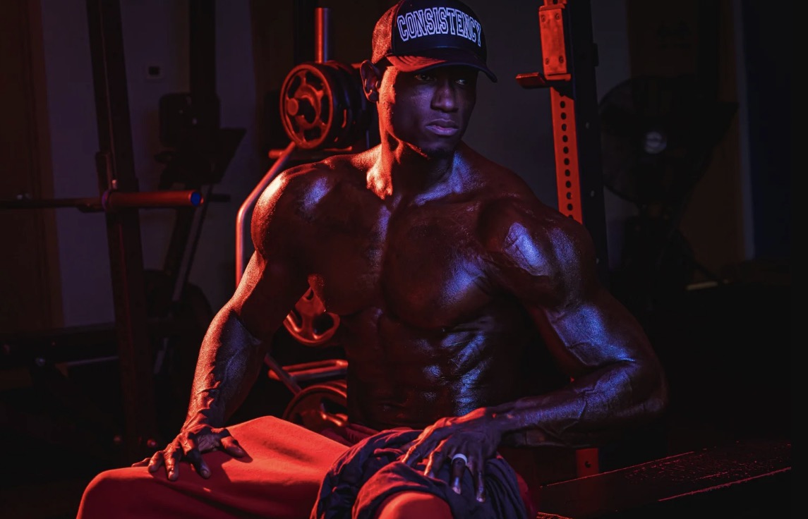 Coach JR seated on a bench in a dark gym wearing a Consistency hat